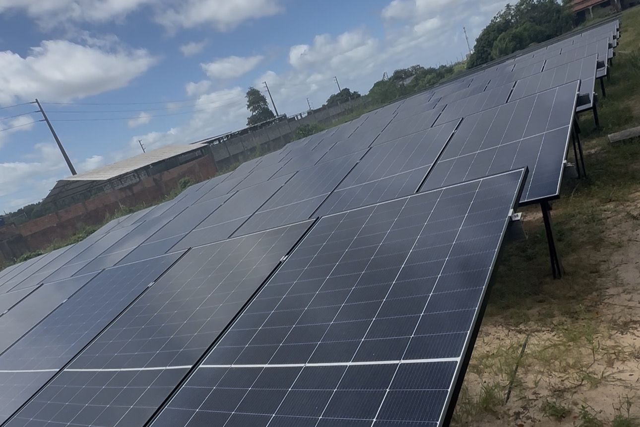 Fileiras de módulos fotovoltaicos em estrutura de solo, sob céu azul com nuvens.